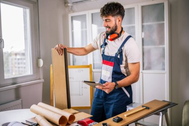 Tamirci apartman zeminini onarıyor, etrafı lamine tahtalarla çevrili, ve rehberlik için bir tablet kullanıyor, modern teknolojiyi ustalıkla birleştiriyor..