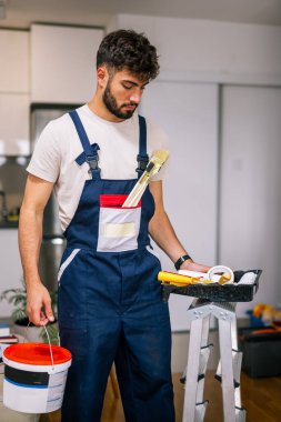 Bir dairedeki ressam, duvarları boyamaya hazırlanıyor, elinde kireç ve fırça tutuyor, yenilenmeye hazır..