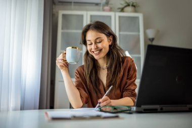 Motive olmuş genç bir kadın ev ortamında üretken ve ilham verici bir şekilde otururken dizüstü bilgisayarı ve kahvesiyle evden çalışıyor..