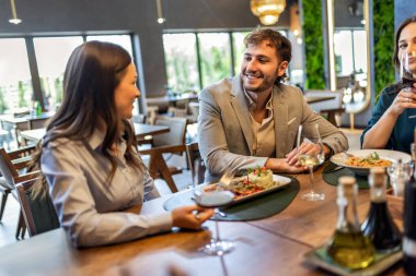 Bir grup iş arkadaşı, pizza, makarna, tavuk, pirinç ve şarapla bir restoranda yemek yerken masada gülümsüyor ve iş hakkında konuşuyor..