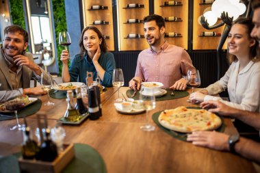 Bir grup iş arkadaşı, pizza, makarna, tavuk, pirinç ve şarapla bir restoranda yemek yerken masada gülümsüyor ve iş hakkında konuşuyor..