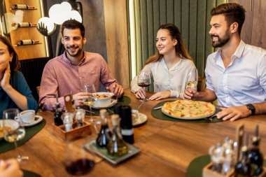 Bir grup iş arkadaşı, pizza, makarna, tavuk, pirinç ve şarapla bir restoranda yemek yerken masada gülümsüyor ve iş hakkında konuşuyor..