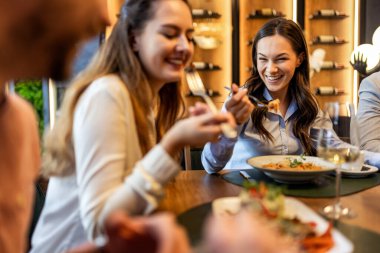 Bir grup iş arkadaşı restoranda öğle yemeği yiyor, kadınlar yemeklerini yerken diğerleri iş hakkında konuşuyor, şarap ve tabaklar masada..