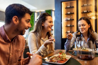 Bir grup iş arkadaşı restoranda öğle yemeği yiyor, kadınlar yemeklerini yerken diğerleri iş hakkında konuşuyor, şarap ve tabaklar masada..