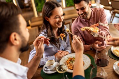 Lokantadaki bir grup arkadaş, büyük miktarda yiyecekle gülümseyen ve eğlenen bir kadına neşe içinde lokma veriyor..