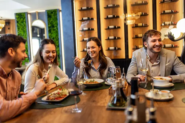 Bir grup iş arkadaşı restoranda öğle yemeği yiyor, kadınlar yemeklerini yerken diğerleri iş hakkında konuşuyor, şarap ve tabaklar masada..