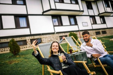 Mutlu genç çift, sonbaharda otelin önünde dinlenirken birlikte selfie çekiyorlar, rahat ve romantik sonbahar anlarının tadını çıkarıyorlar..