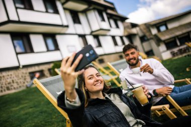 Mutlu genç çift, sonbaharda otelin önünde dinlenirken birlikte selfie çekiyorlar, rahat ve romantik sonbahar anlarının tadını çıkarıyorlar..