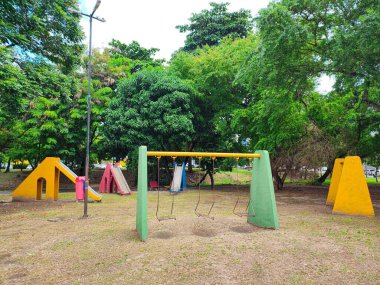 Yeşil bir alanda, renkli oyun alanı ekipmanlarıyla dolu bir park görüyoruz, hepsi boş, çocuksuz bir parkın yalnızlığını vurguluyor..