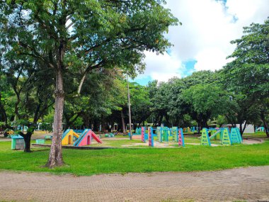 Renkli mavi, pembe ve yeşil ekipmanlarla dolu canlı bir oyun alanı, yemyeşil ağaçlarla çevrili ve parçalı bulutlu gökyüzü, çocuklar için neşeli ve davetkar bir atmosfer yaratıyor..