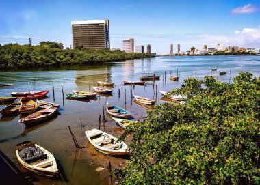 Mangrovlarla kaplı geniş bir nehrin alçak sularında, şehir binalarıyla demirli ahşap tekneler arasındaki zıtlığı görüyoruz..