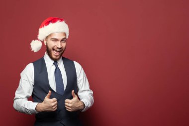 Takım elbiseli ve Noel şapkalı neşeli başarılı iş adamı kırmızı stüdyo arka planında baş parmağını gösteriyor.