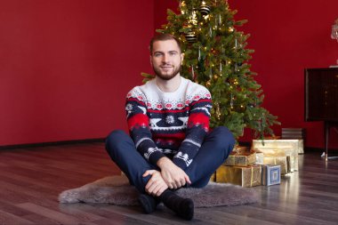 Olumlu yakışıklı adam Noel ağacının yanında oturuyor ve Noel Baba 'nın hediye kutusunu paketliyor. 