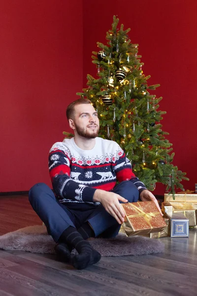 Cute man in sweater sits by nice Christmas tree and holds his gold gift