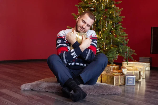 Cute man in sweater sits by nice Christmas tree and feels happy about his gold present