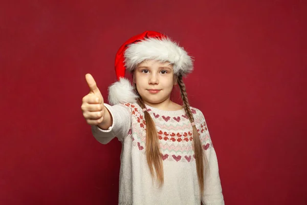 かわいいです子供女の子でサンタキャップ保持親指上の赤いバナーの背景 クリスマスの肖像画 — ストック写真