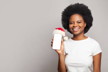 Beyaz tişörtlü tatlı Afro-Amerikan kadın portresi gri arka planda Noel Baba şapkalı akıllı telefon.