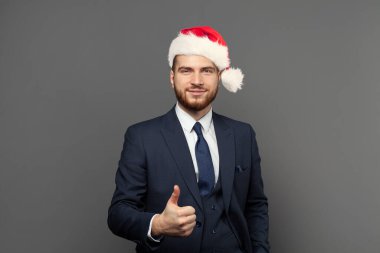 Noel şapkalı akıllı iş adamı gri duvar arka planında baş parmağını kaldırıyor. İş zaferi, Noel ve başarı kavramı