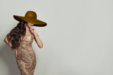 Uzun kıvırcık saçlı, altın rengi elbise giyen ve beyaz arka planda geniş kahverengi geniş kenarlı şapka takan mutlu bir genç kadın.