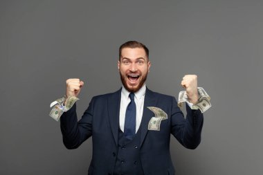 Takım elbiseli yakışıklı iş adamı çığlık atıyor ve cebinde dolarlarla gülüyor gri stüdyo arka planında. İş kazanma, para ve başarı konsepti