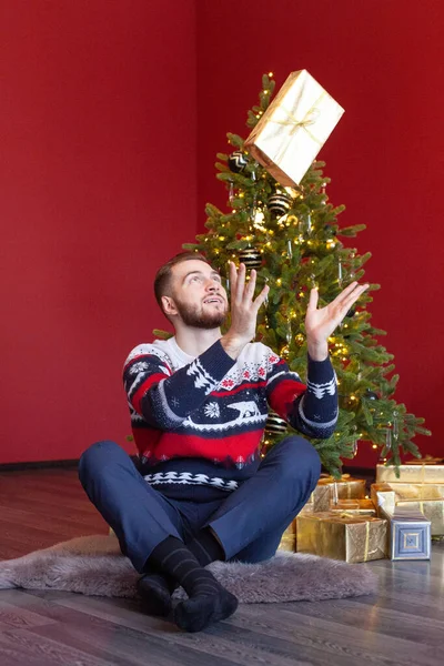 Cute man in sweater sits by nice Christmas tree and throws up his gold Santa gift on the red background