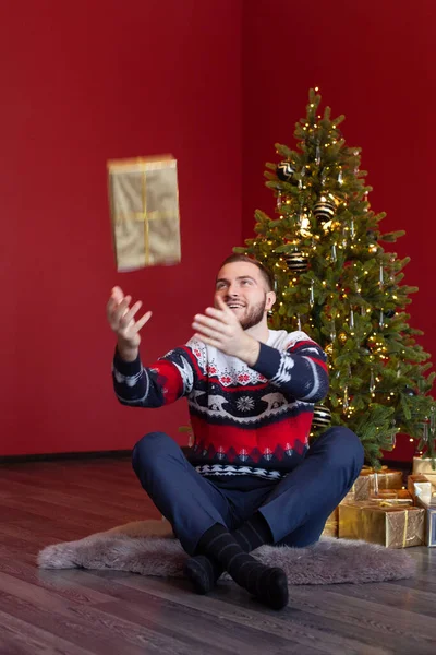 Happy man in sweater sits by Christmas tree and throws up his gold santa claus gift