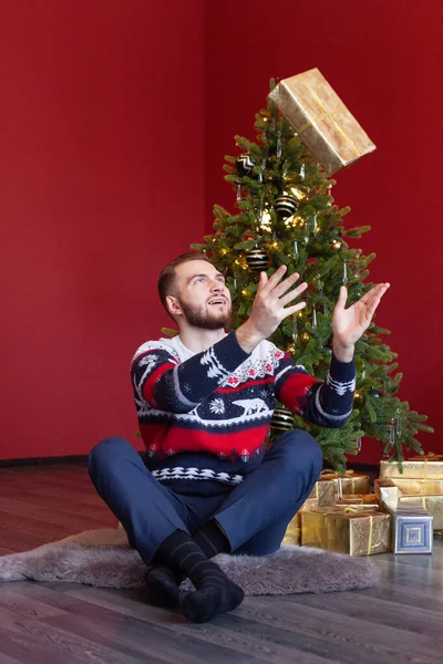 Cute man in sweater sits by nice Christmas tree and throws up his gold Santa gift