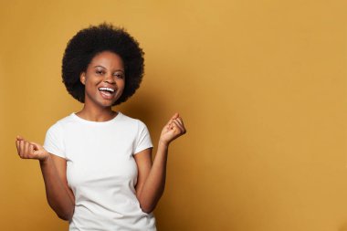 Beyaz tişörtlü neşeli bir kadın model. Parlak sarı stüdyo duvarına gülüyor ve eğleniyor.