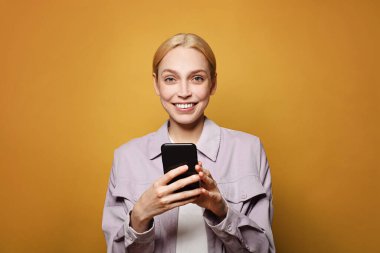 Mutlu bir kadın, sarı renkli bir stüdyo arka planında telefonla mesajlaşıyor.