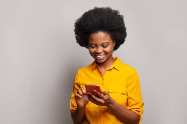 Sarı tişörtlü, beyaz stüdyo duvarına karşı akıllı telefon kullanan esmer bir kadın.