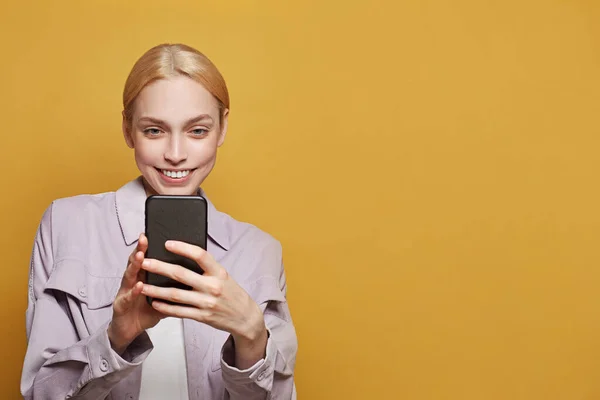 Young pretty woman using app or sending a message with the mobile phone on colorful yellow background