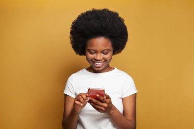 Pretty woman in white t-shirt using smart phone and enjoying apps against yellow studio wall banner background