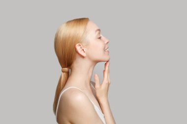 Fresh blonde woman in white top standing sideways isolated on grey background. Beauty, cosmetics, skincare, glamour