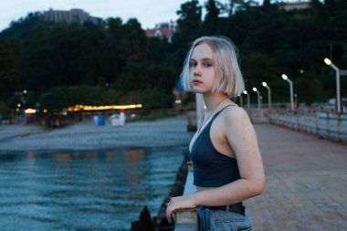 Mavi kolsuz tişörtlü genç sarışın kadın mavi denizin ve akşam plaj parkının arka planında duruyor.