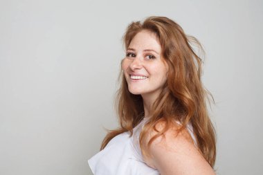 Mutlu kızıl saçlı kadın gülümsüyor ve beyaz arka planda kameraya bakıyor.