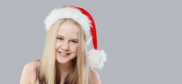 灰色のスタジオの壁のバナーの背景に対して赤いサンタの帽子をかぶっているクリスマスの子供の少女 — ストック写真