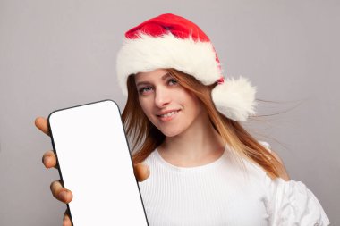 Kırmızı Noel Baba şapkalı kızıl kadın yeni uygulama için beyaz ekranlı akıllı telefonu gösteriyor, Mockup pankartı.