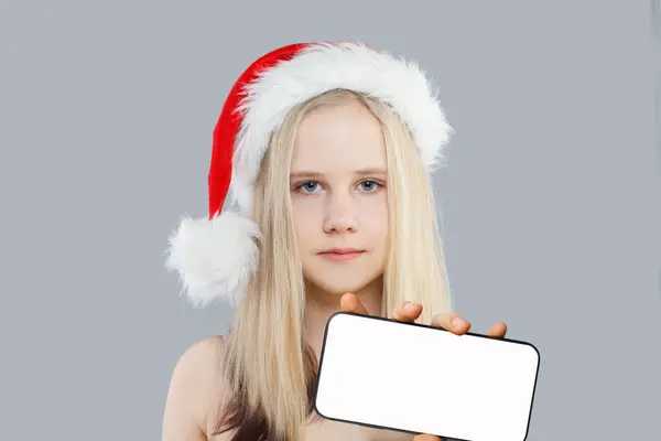 グレーの背景に大きな空のディスプレイを持つスマートフォンを示すクリスマスサンタの少女 — ストック写真