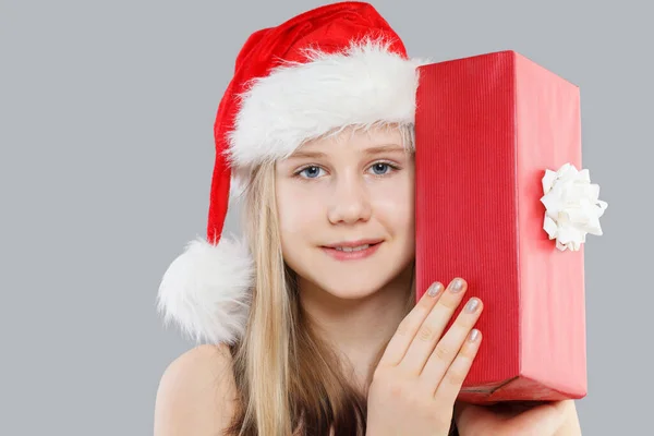 赤いサンタの帽子でかわいい笑顔の少女のクリスマスの肖像画は灰色の背景にプレゼントボックスを保持しています — ストック写真
