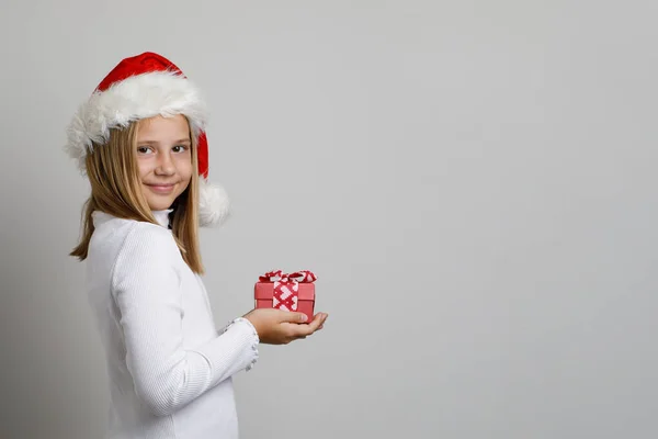 白いスタジオの壁のバナーの背景に対して立っている赤いXmasギフトが付いているクリスマスの子供の少女サンタ — ストック写真