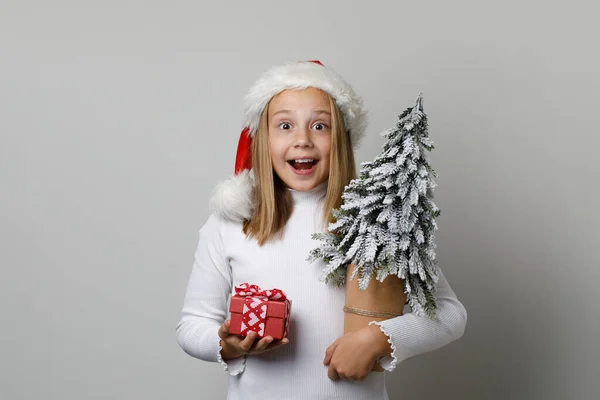 サンタハットの幸せな喜びに興奮する子供の少女は 白い背景にクリスマスツリーと赤いギフトで帽子をかぶります クリスマスコンセプト — ストック写真