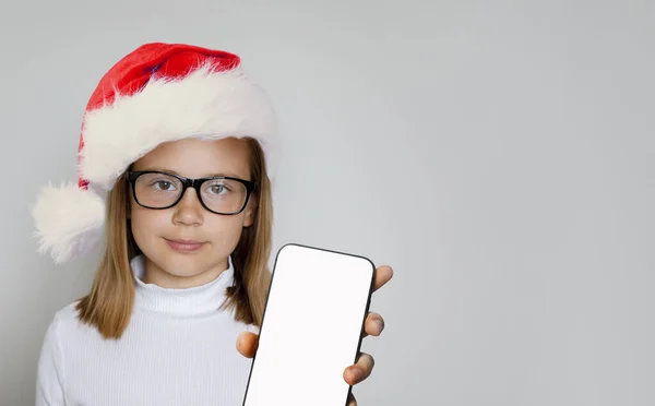 サンタハットの若いスマートな女の子は空白の白い画面の表示が付いているスマートフォンを示します クリスマスコンセプト — ストック写真