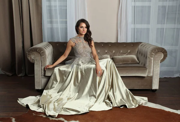 迷人的女人穿着时髦的丝质闪亮的银色连衣裙在褐色的沙发上悠闲自在 时尚的美丽肖像 — 图库照片