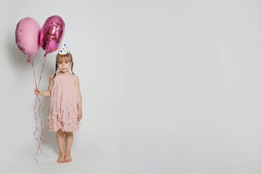 Beyaz stüdyo duvarında iki pembe balon tutan neşeli çocuk. Doğum günün kutlu olsun.