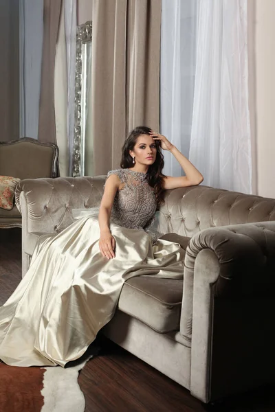 一个年轻貌美的黑发女子穿着闪闪发亮的银色连衣裙坐在室内沙发上的画像 — 图库照片
