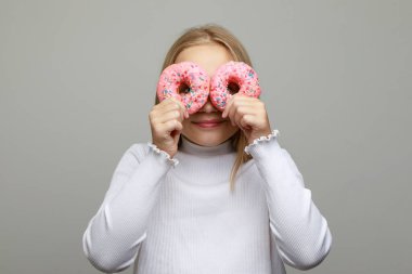 Gri arka planda gözlerini pembe ve çikolatalı çöreklerin arkasına saklayan tatlı bir kız çocuğu. Doğum günü partisi konsepti