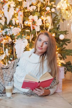 Genç çekici esmer kadın evde Noel ağacının önünde kitap okuyor.