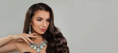 Uzun dalgalı saç modeli, gri arka planda elmas küpe ve kolyesi olan çekici esmer kadın, moda güzellik portresi.