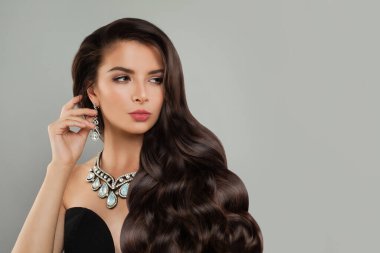 Uzun kıvırcık saçlı, şık model kadın esmer, gri arka planda elmas küpe ve kolye, moda güzellik portresi.