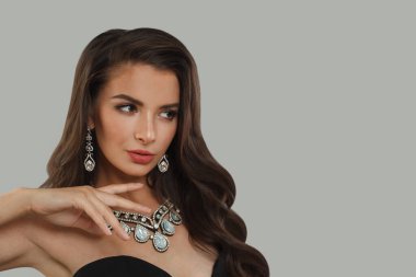 Koyu kahverengi saçlı, parlak tenli, pırlanta küpeli, kolyeli muhteşem bir mücevher mankeni. Gri stüdyo duvarına poz veriyor. Moda güzelliği portresi.